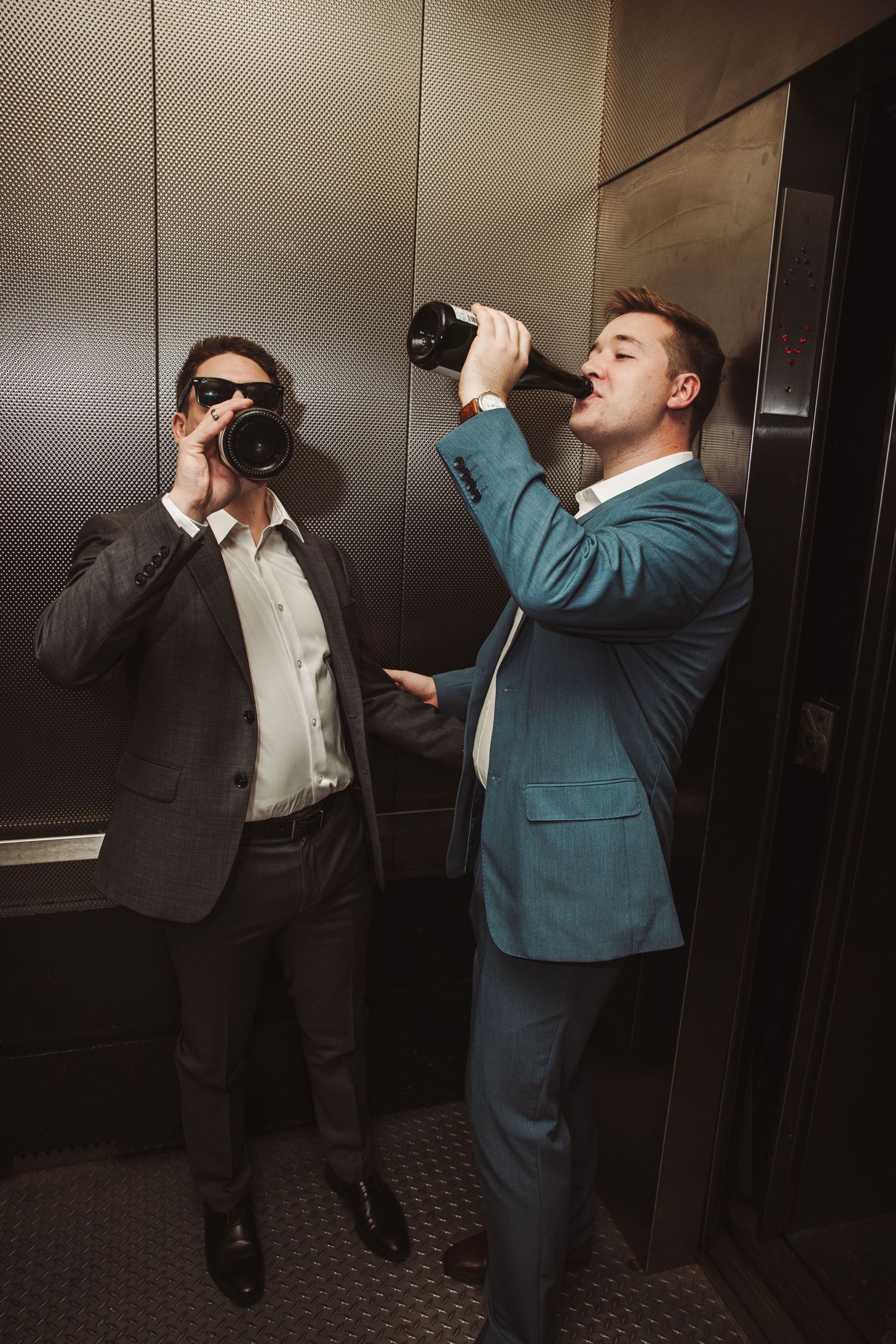 Two men in suits drinking from wine bottles in an elevator at a wedding.