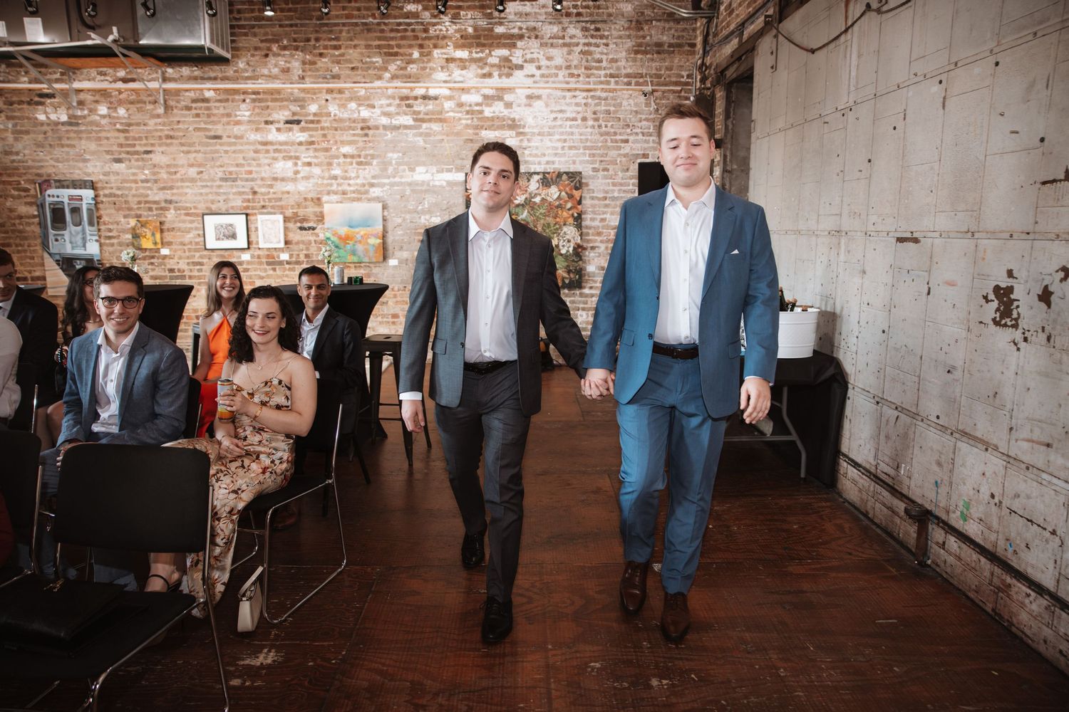 Two grooms in suits walking hand in hand down aisle after wedding ceremony.