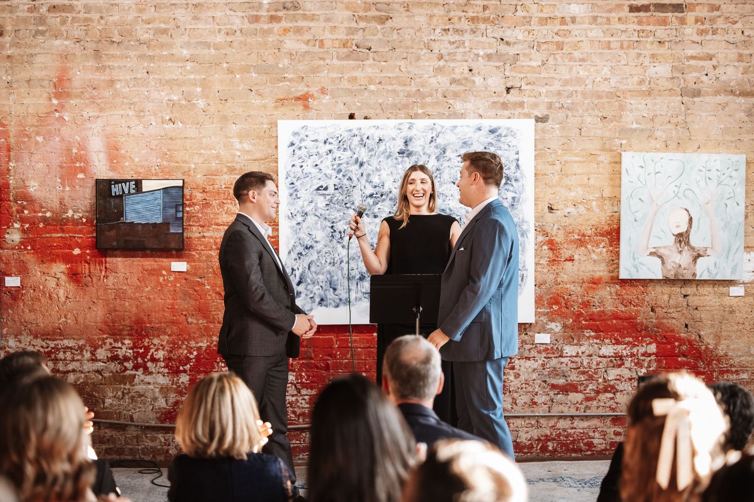 Woman at podium facilitates a handshake between two men at a formal indoor ceremony.