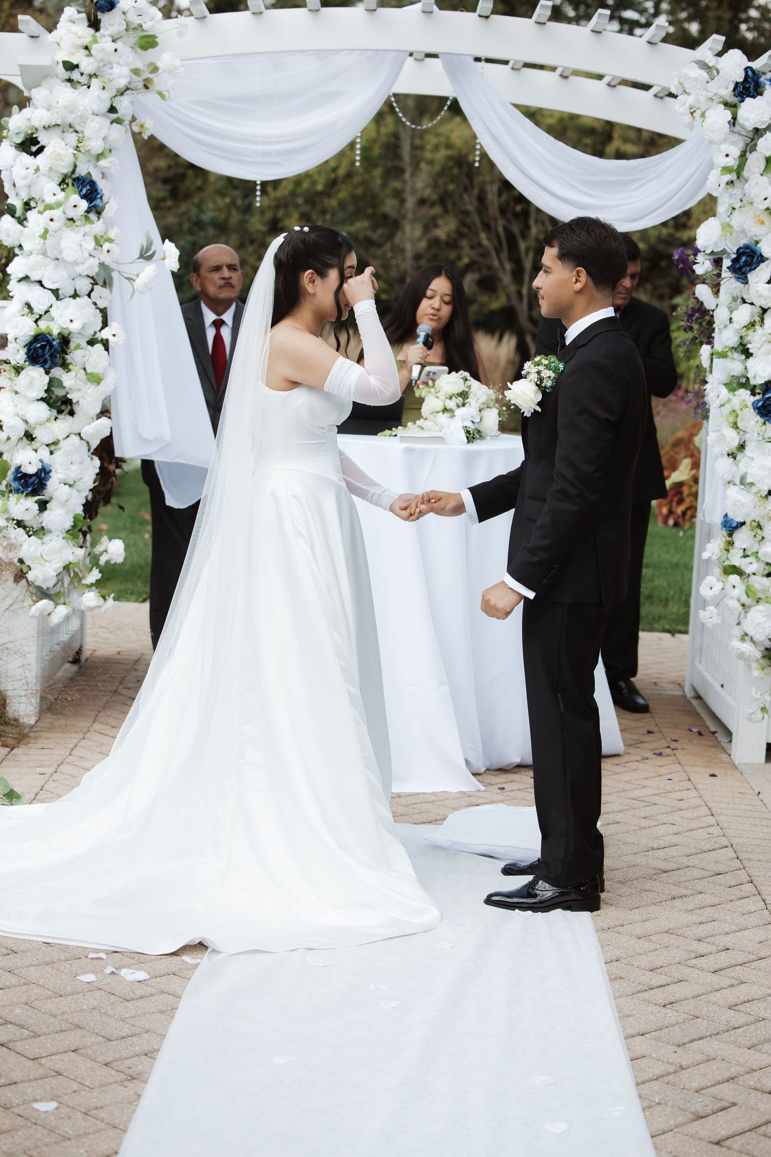 Emotional bride wipes tears as groom holds her hand at outdoor wedding ceremony.