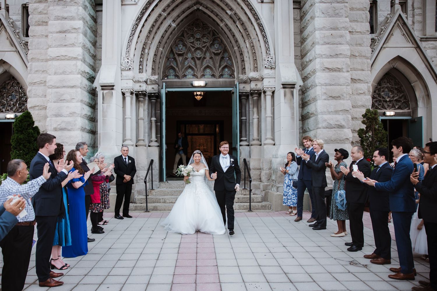 Joyful newlywed couple walks through crowd of celebrating guests outside majestic gothic stone church.