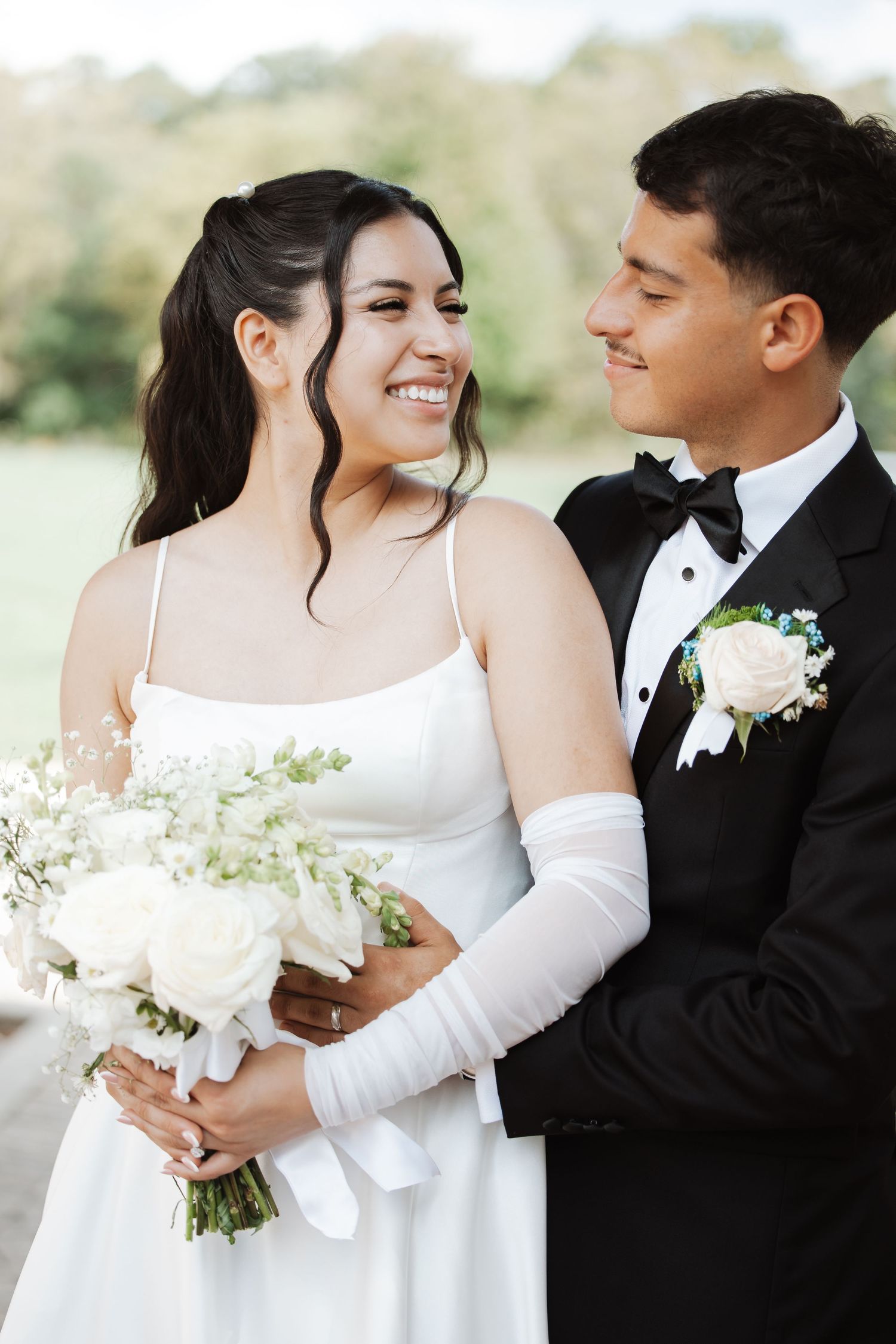 Happy bride and groom smiling at each other outdoors, bride holding white rose bouquet.
