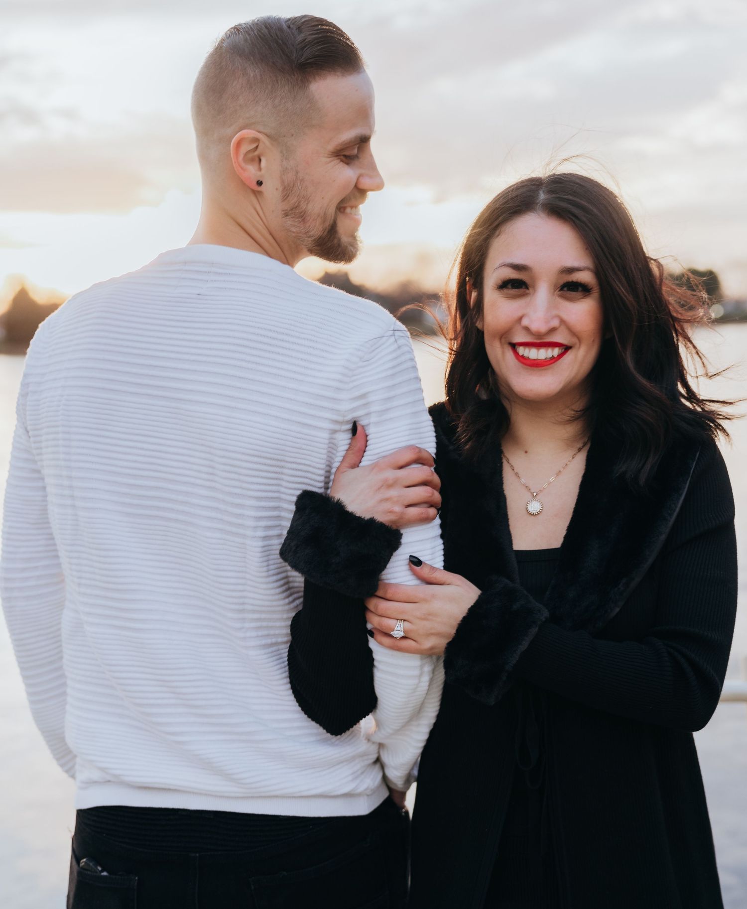 Couple sharing a romantic moment at sunset by the water, woman in black coat smiling brightly.