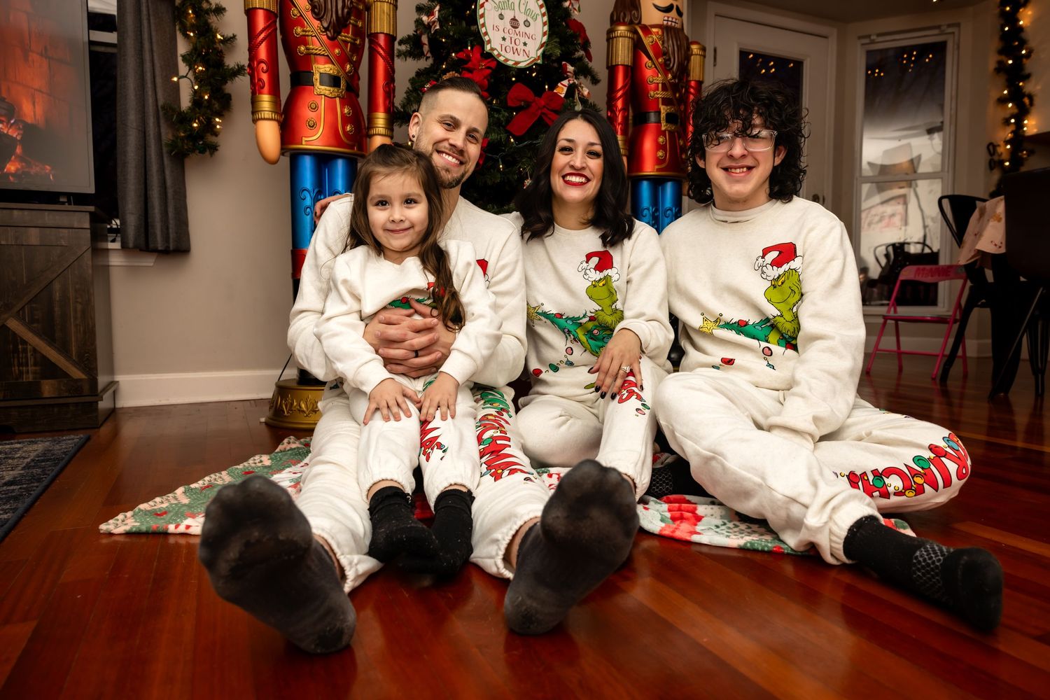 Smiling family of four wearing matching white Grinch holiday pajamas posing by Christmas tree.