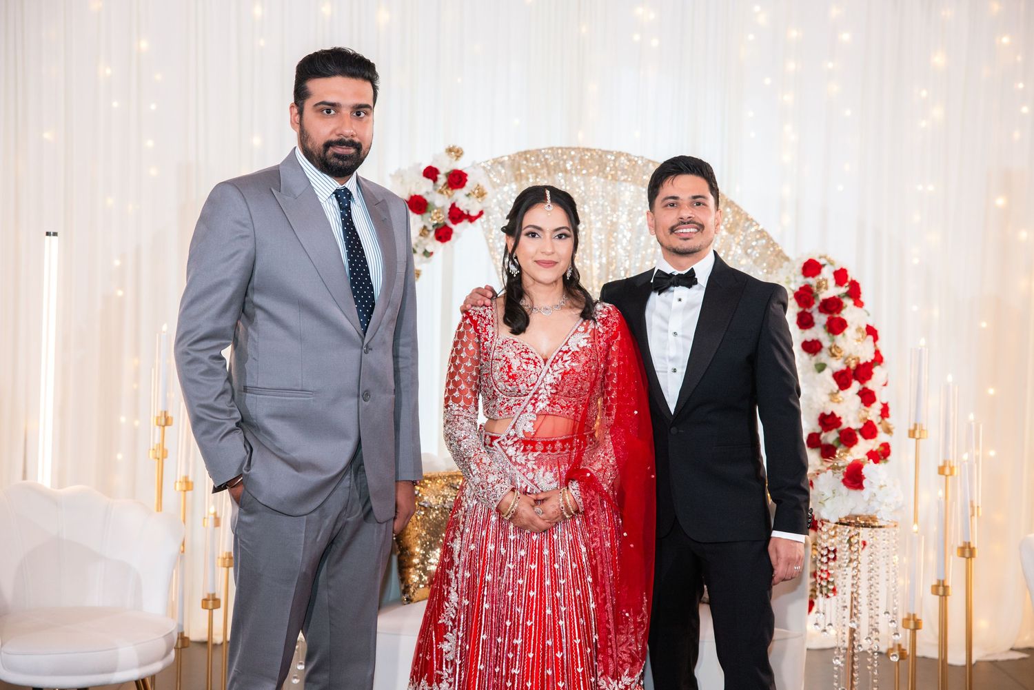 Bride in red lehenga poses with two men in formal suits at decorated Indian wedding reception.