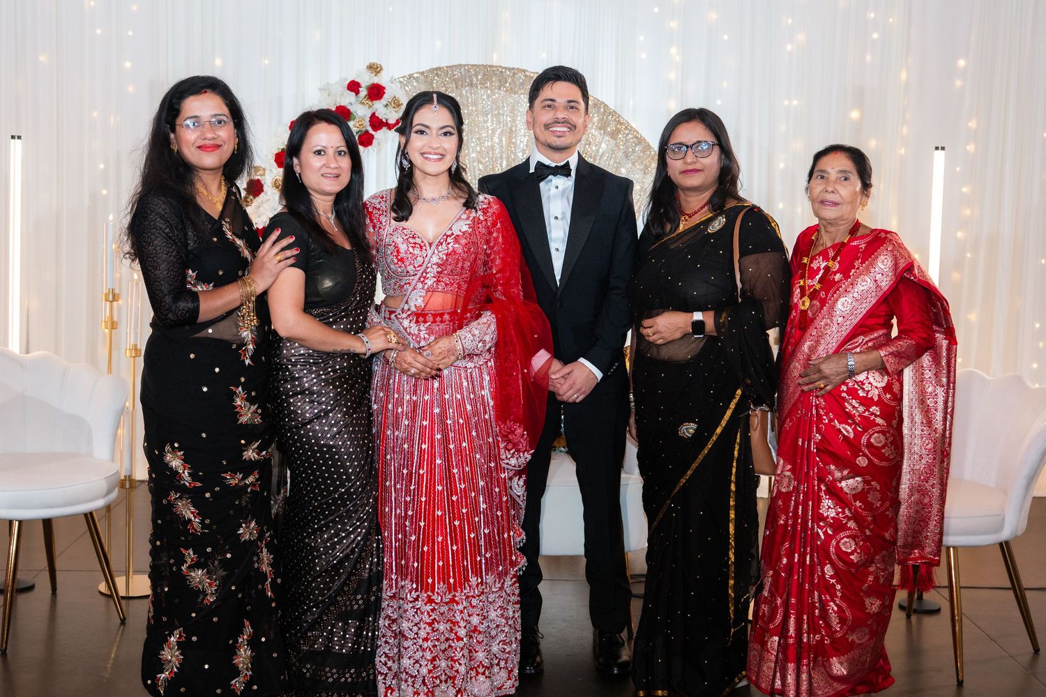 Bride in red lehenga with groom and four women in traditional black and red sarees at wedding.
