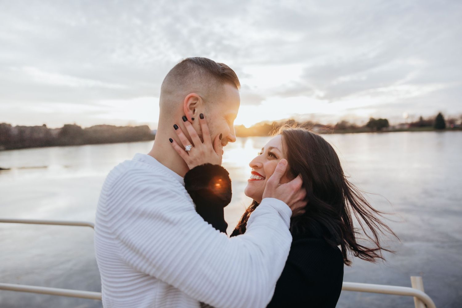 Couple almost kissing at golden hour by a lake, woman showing engagement ring while man cradles her face tenderly.