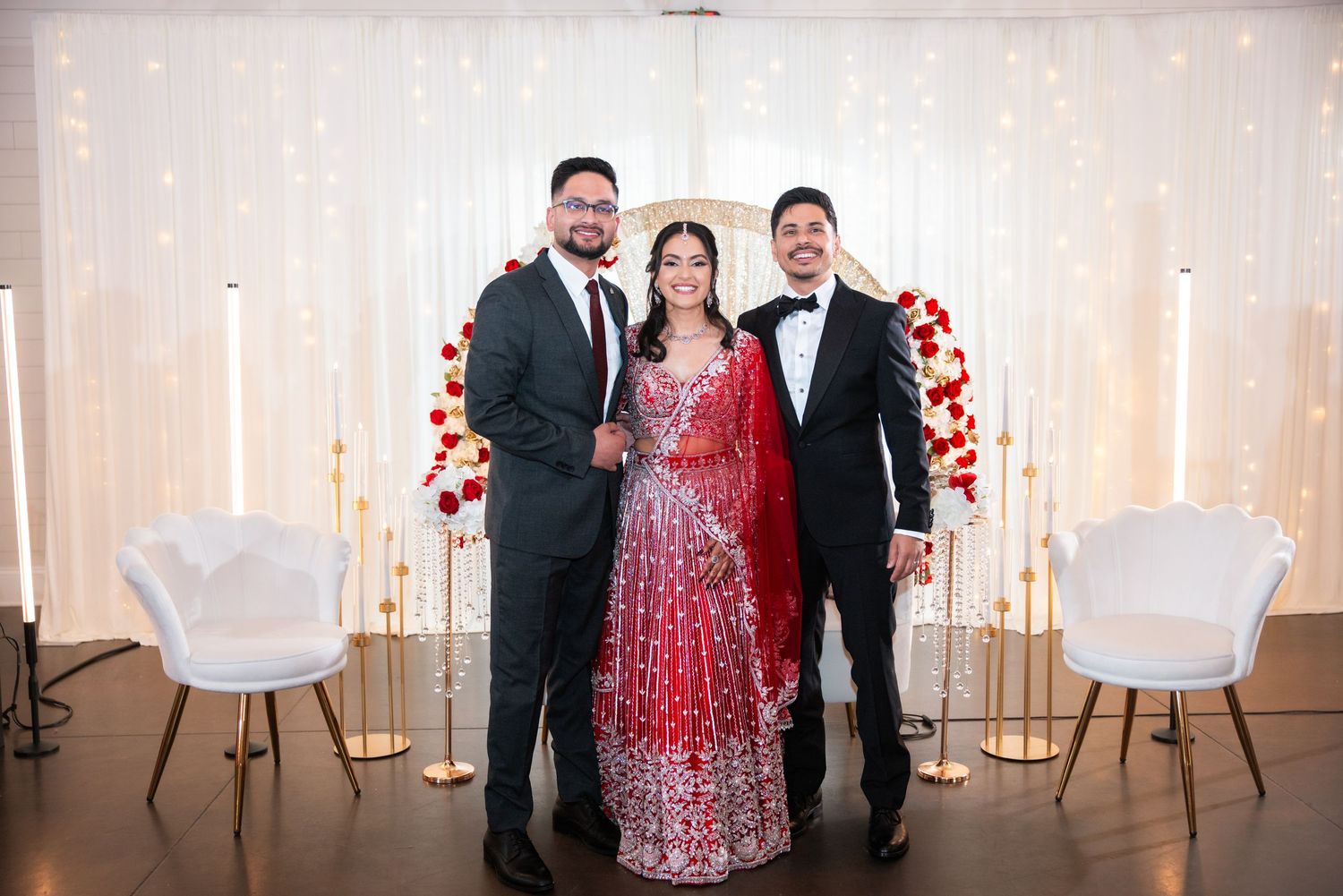 Indian bride in red bridal lehenga poses with two men in suits at wedding reception.