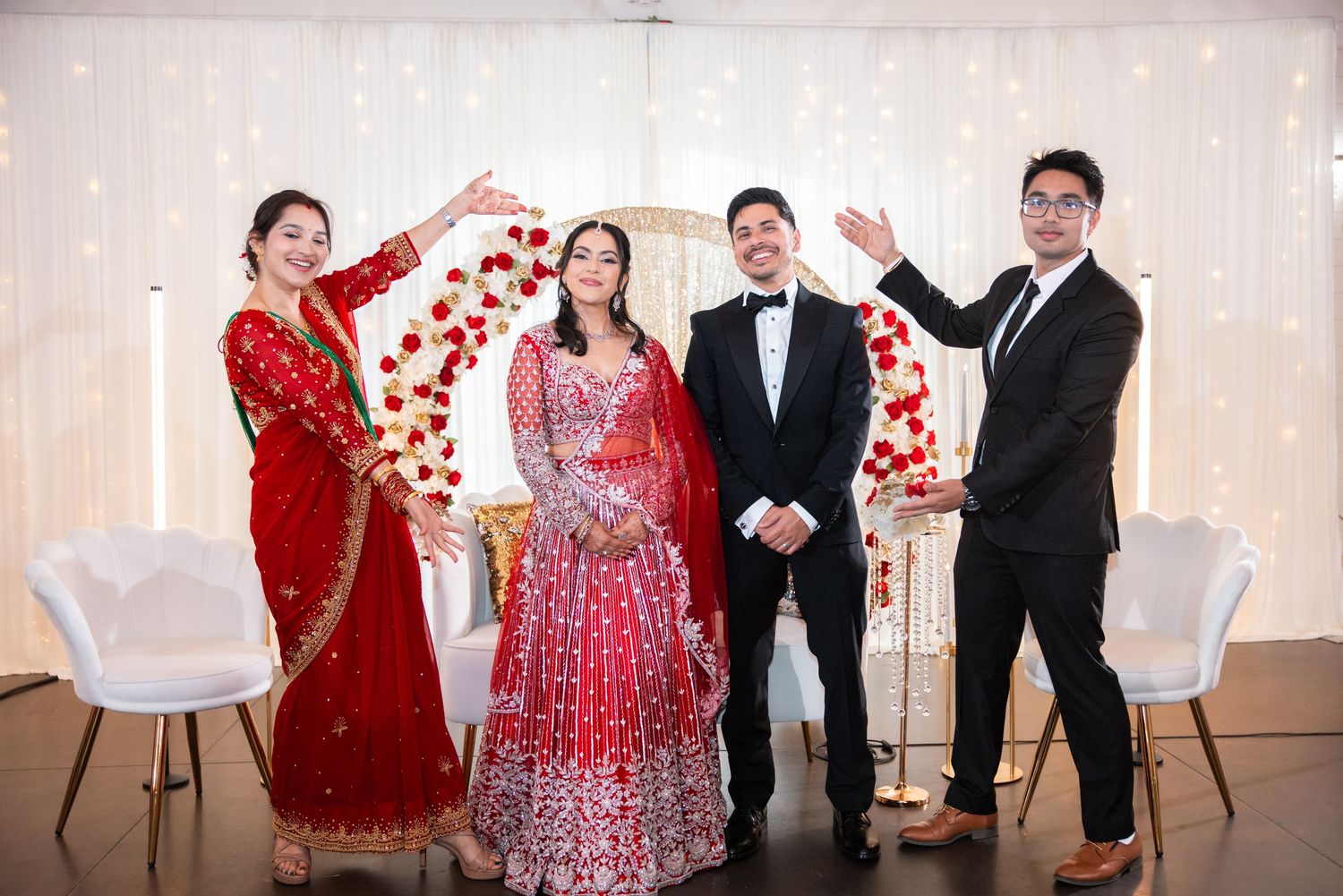 Indian bride and groom in tuxedo pose with two guests making arch gesture at wedding.