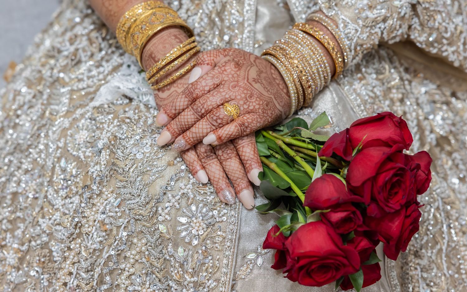 Bride's henna-adorned hands with gold bangles hold a bouquet of red roses in embroidered wedding dress.
