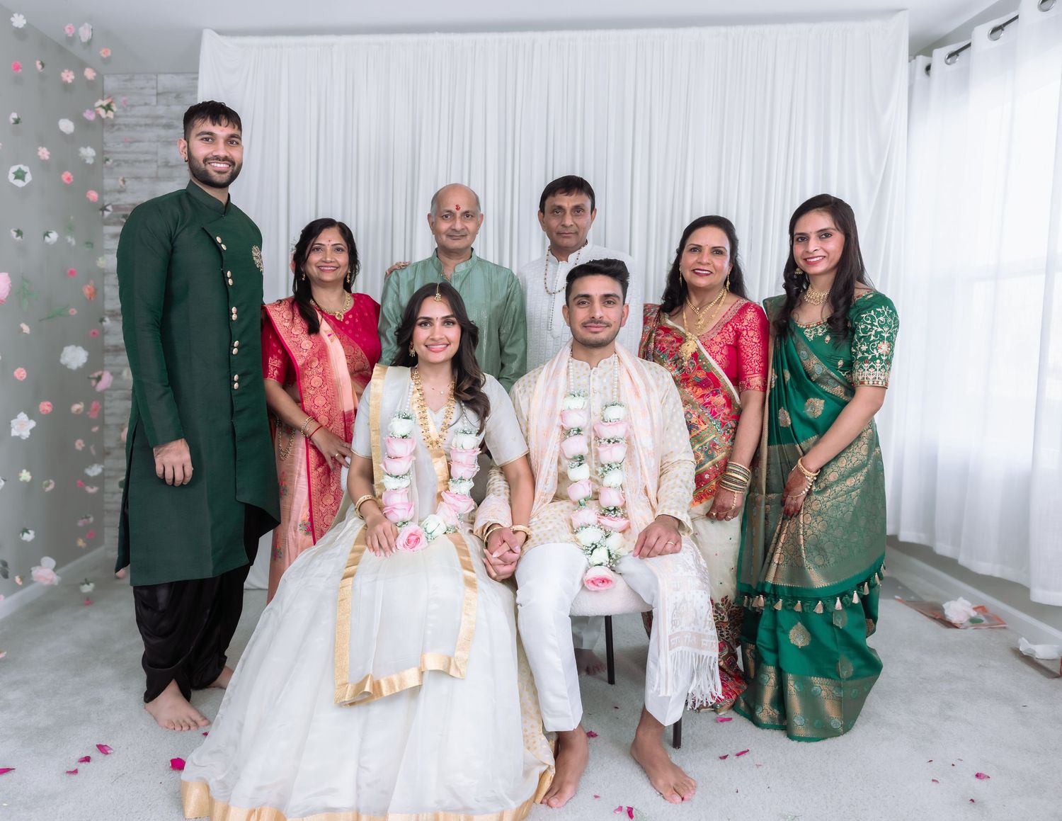 Indian wedding family group photo with bride in white lehenga and groom in sherwani with floral garlands.
