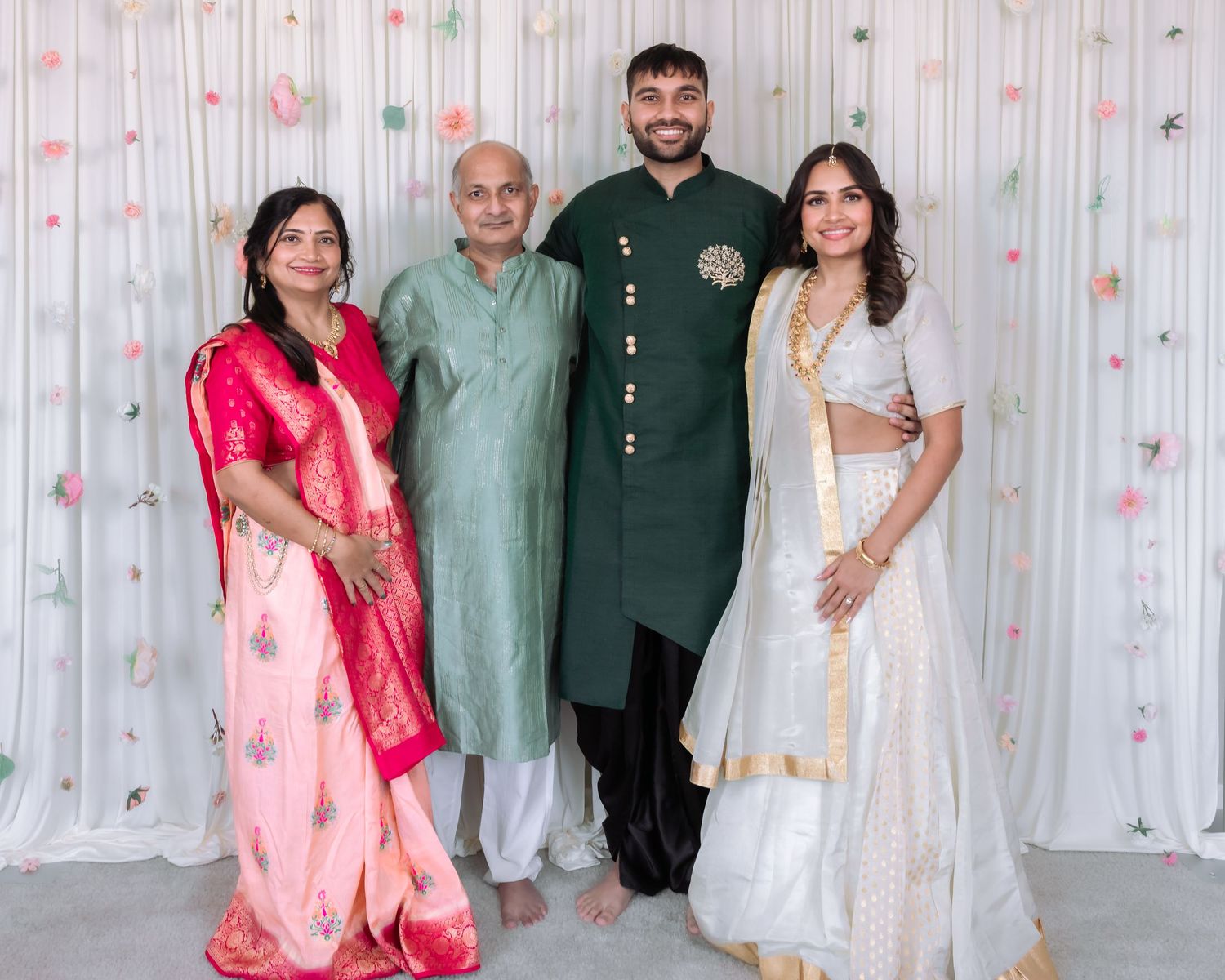 Indian family of four in traditional attire pose together at wedding celebration with floral decorated white backdrop.
