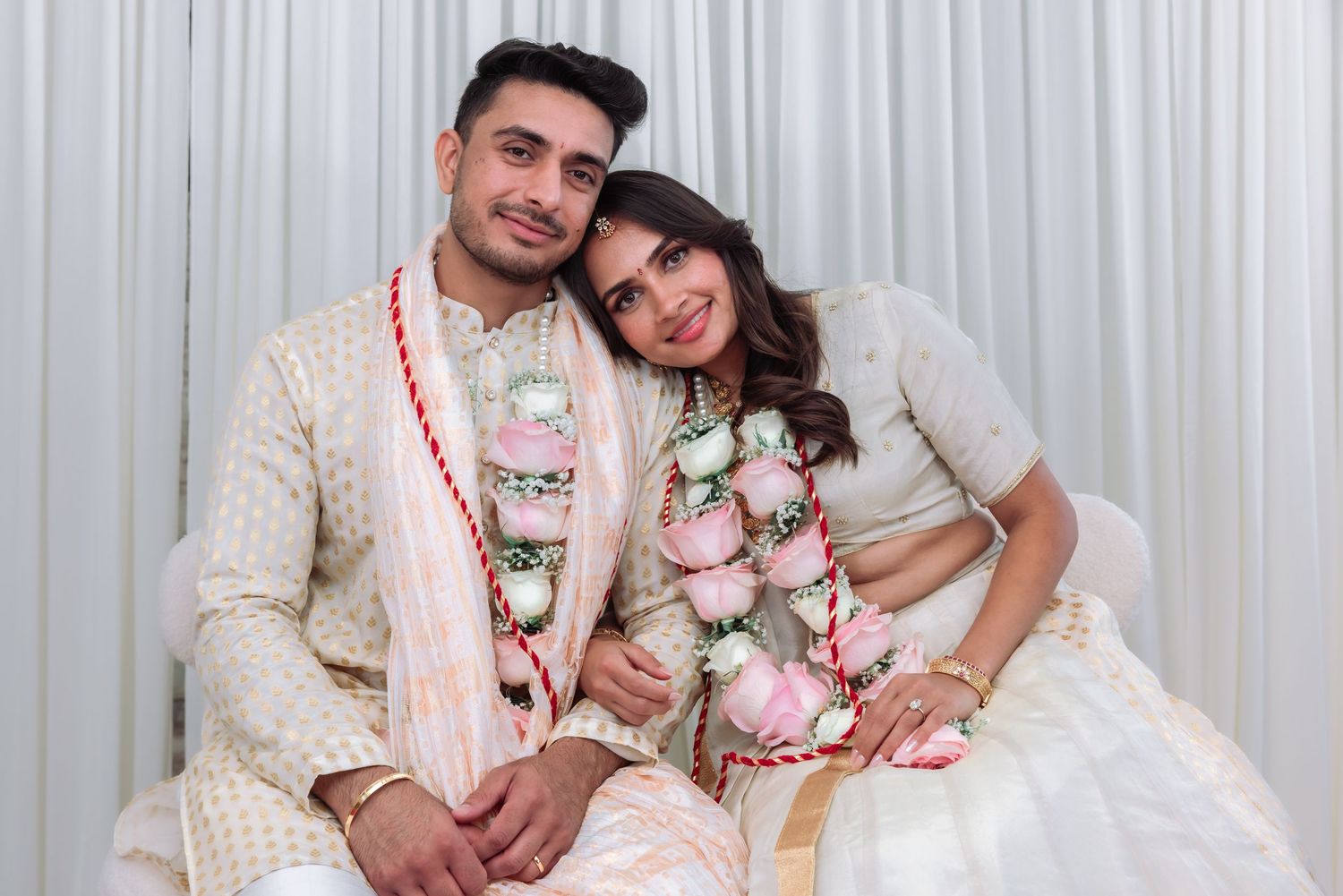 Indian couple in traditional white and cream wedding attire posing with floral garlands at engagement ceremony.
