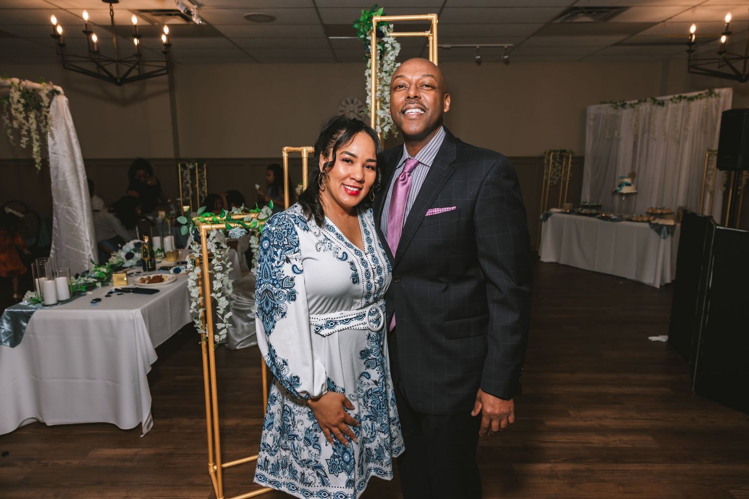 Couple posing together at a wedding reception, man in suit and woman in blue floral dress.