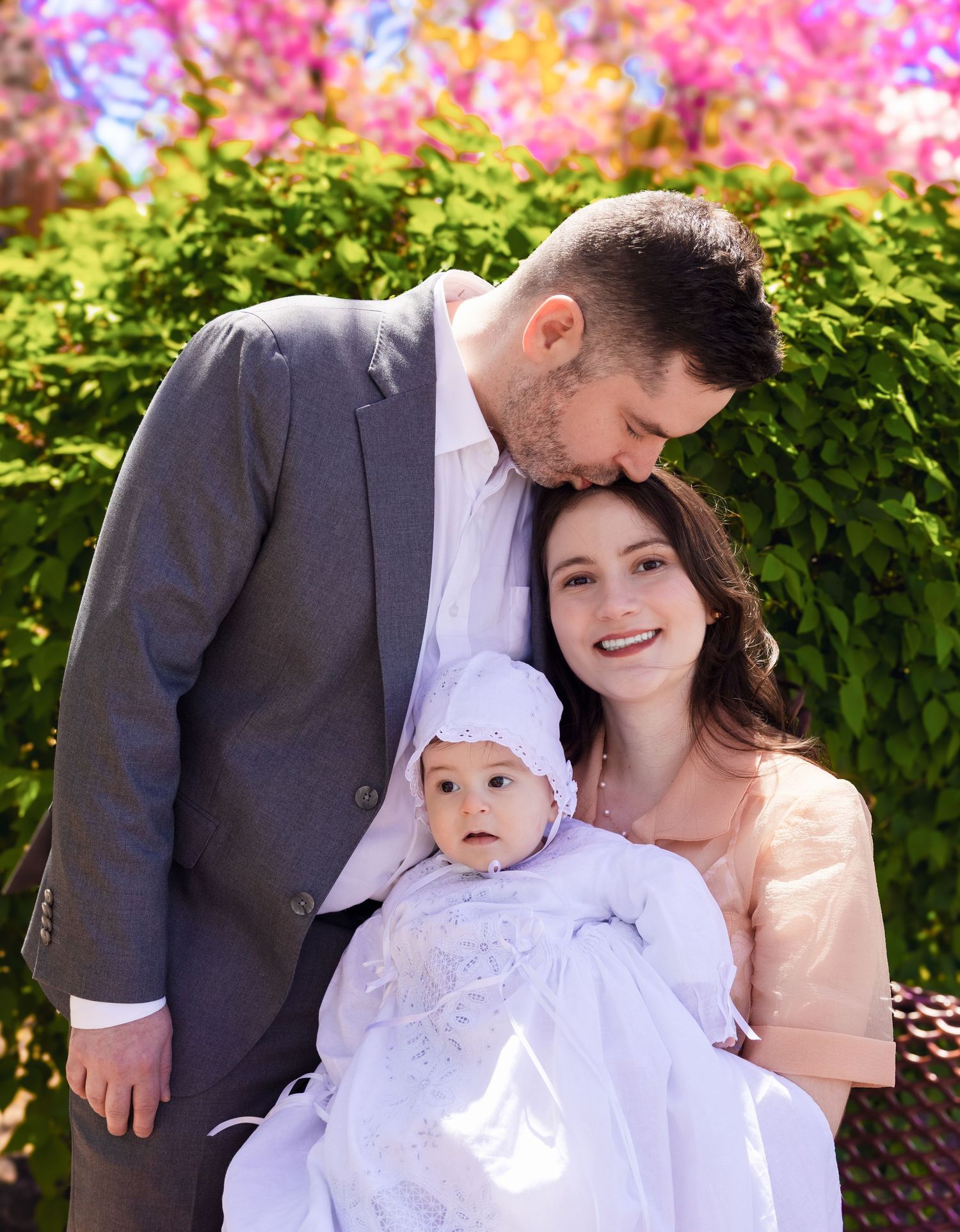 Couple kissing baby dressed in white christening gown with pink flowering trees in background.