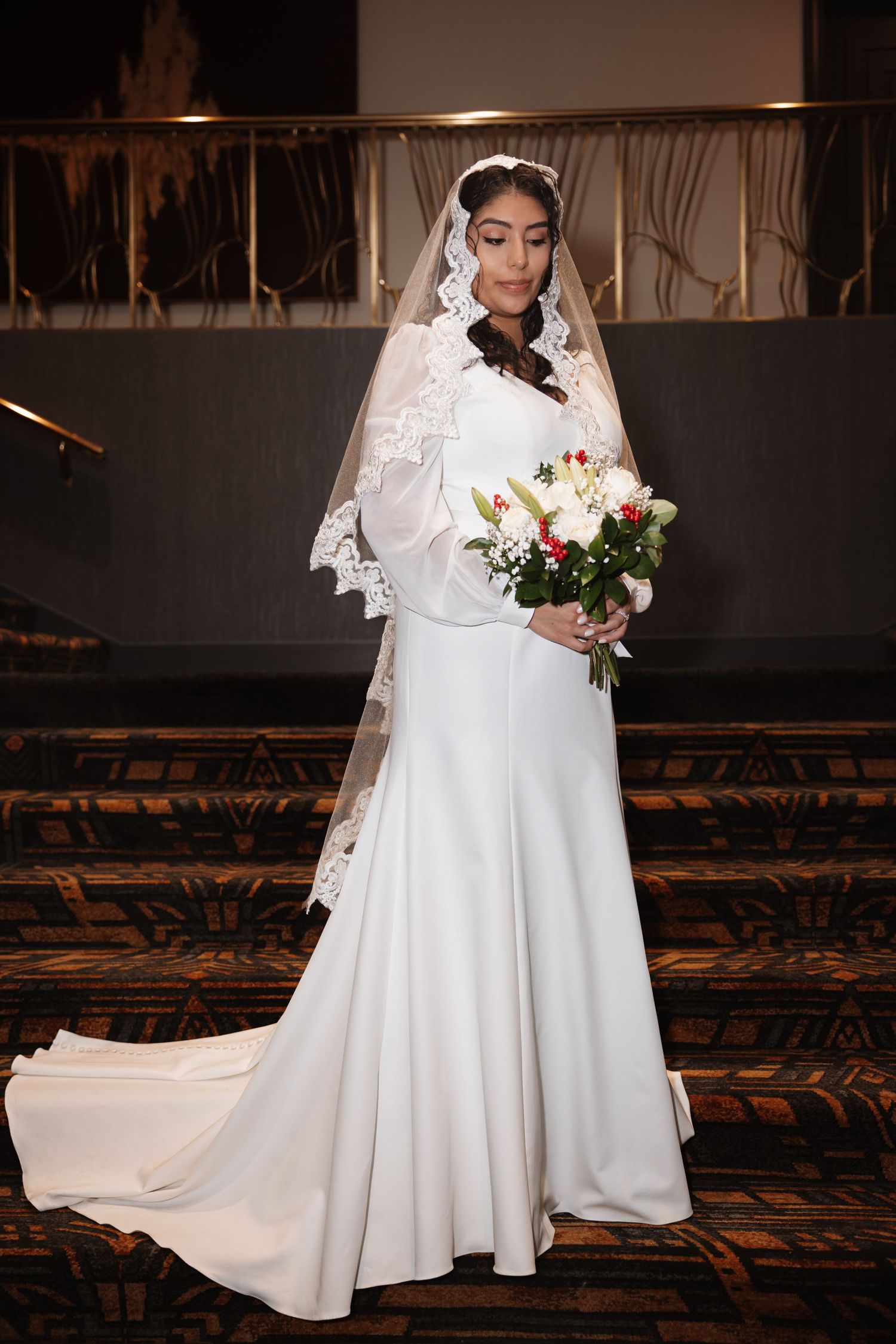 Elegant bride in white fitted gown and lace veil holds floral bouquet while looking down near staircase.