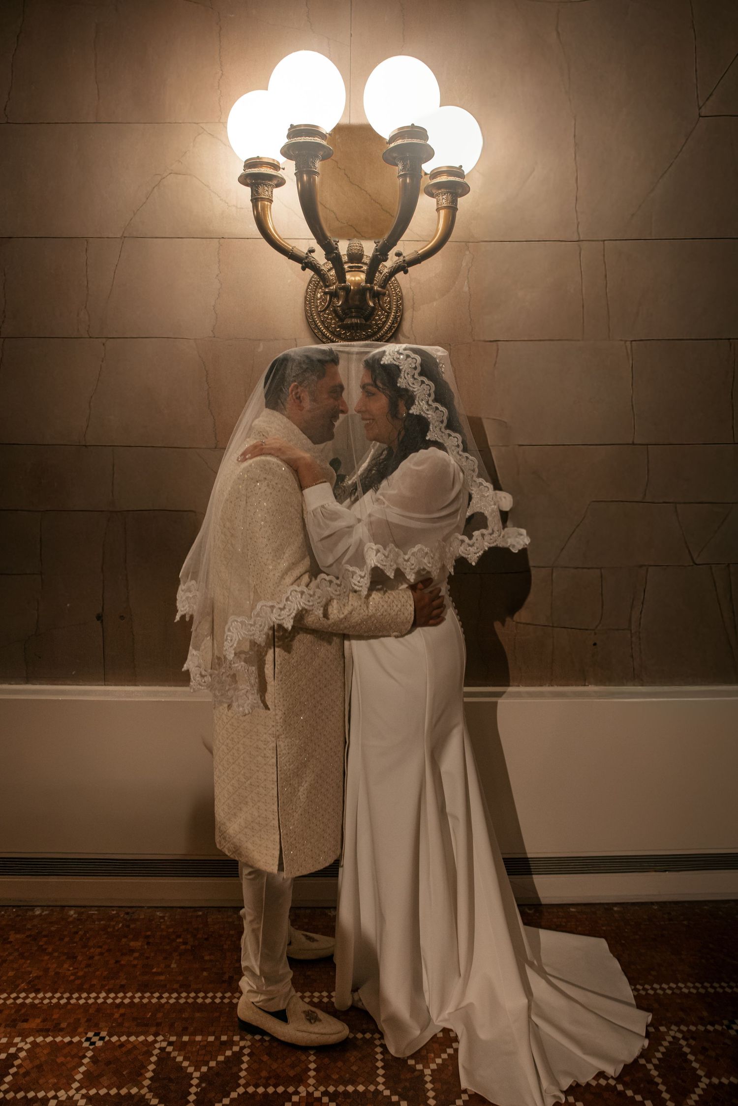 Wedding couple embracing and smiling at each other beneath an elegant lit candelabra wall sconce in a warmly lit venue.