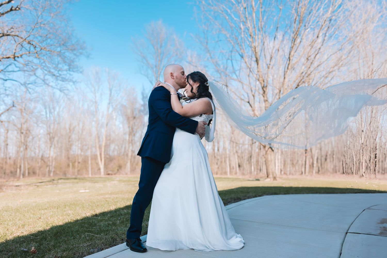 Bride and groom embrace and kiss outdoors with flowing veil under bright blue sky.