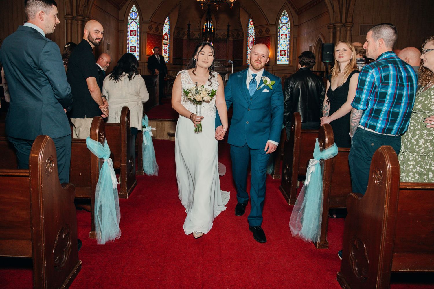 Newlyweds walk hand in hand down the aisle past smiling guests in a church decorated with teal tulle bows.