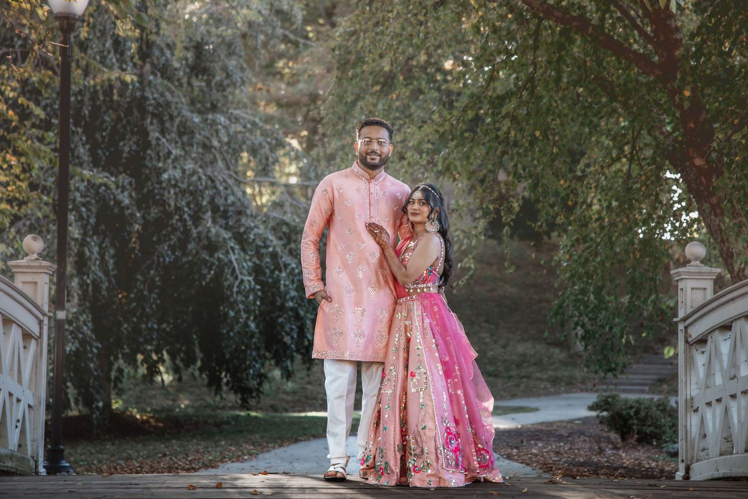 Indian couple in pink traditional wedding attire walks on bridge surrounded by trees.