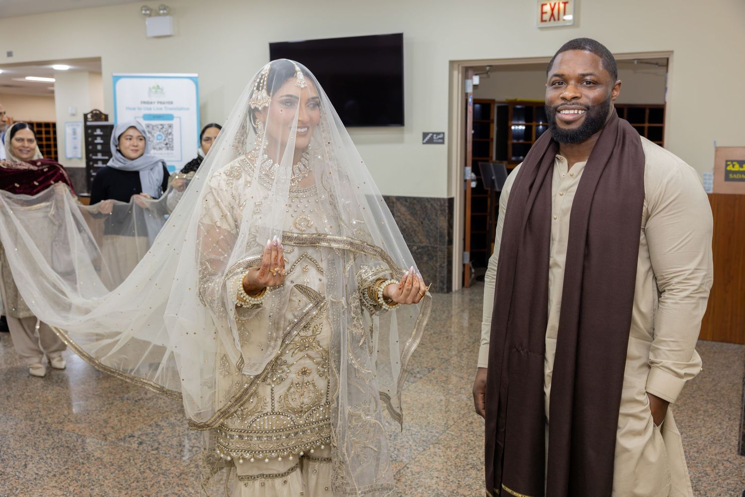A smiling groom in cream outfit stands beside his veiled South Asian bride in a decorated hall.