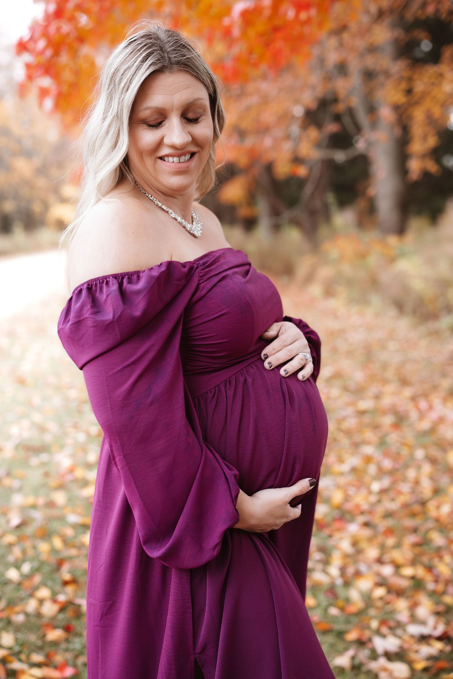 Smiling pregnant woman in purple gown cradling bump outdoors with vivid autumn trees in background.