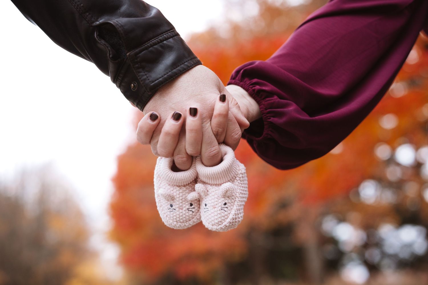 Couple holding hands and white knit baby booties against vibrant autumn foliage background.
