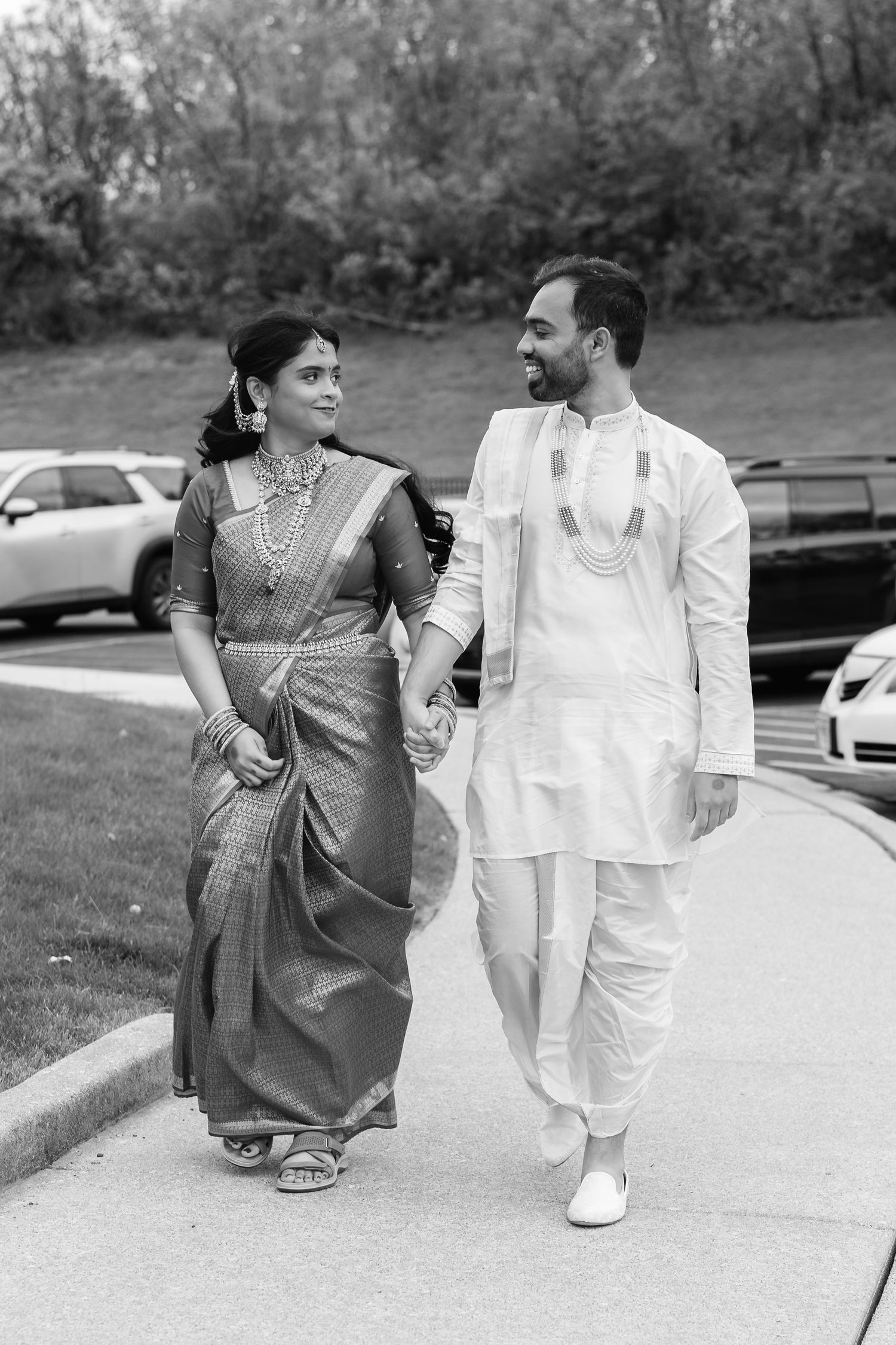 Indian couple in traditional attire holding hands and walking together in a black and white wedding photo.