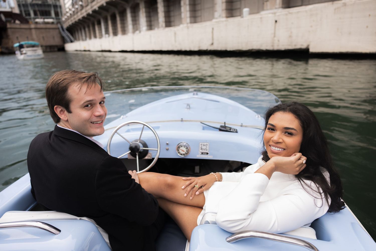 Couple smiling and relaxing together on a light blue vintage boat on calm water near a bridge.
