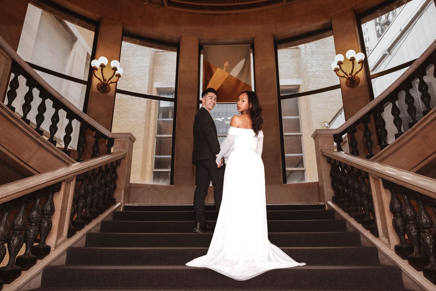 Bride in white gown and groom holding hands on grand curved staircase with warm lighting.Chicago Wedding Photographer