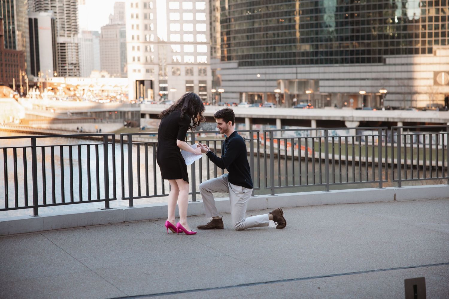 Man on one knee proposing to woman with pink heels on urban bridge with city skyline.
