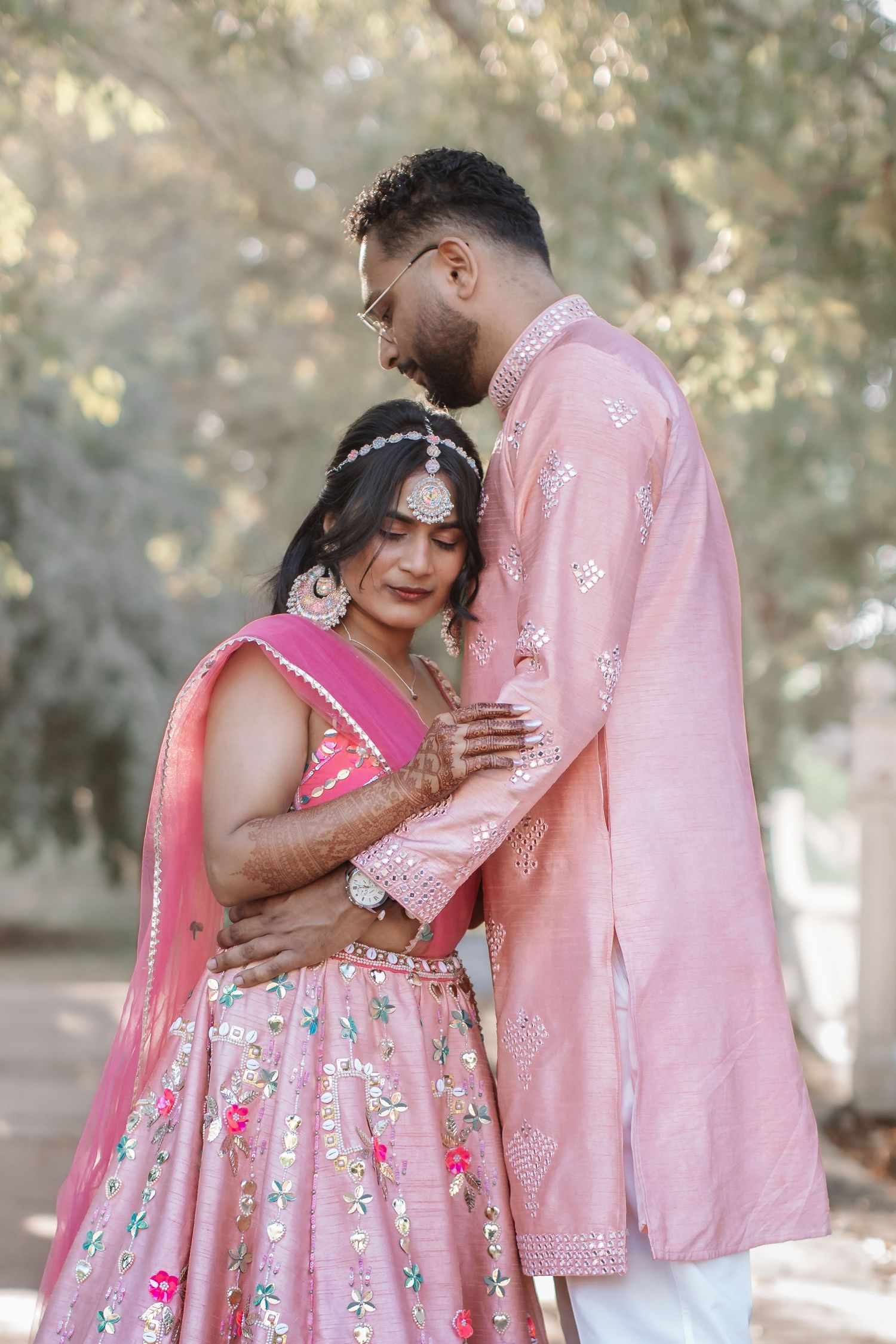 Indian bride and groom in pink traditional outfits sharing a tender moment outdoors.