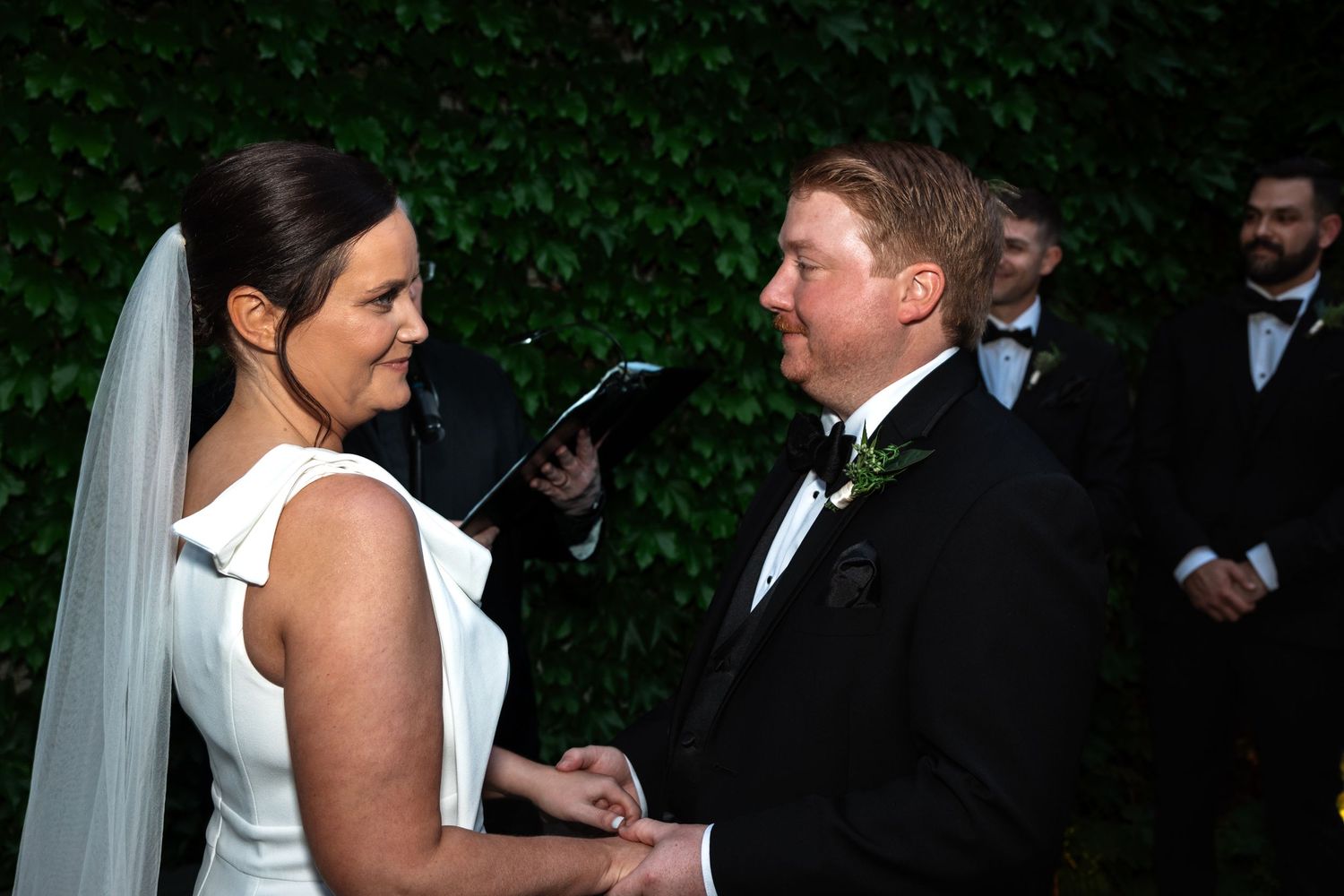 Bride and groom exchange vows holding hands at outdoor wedding ceremony with ivy wall backdrop.