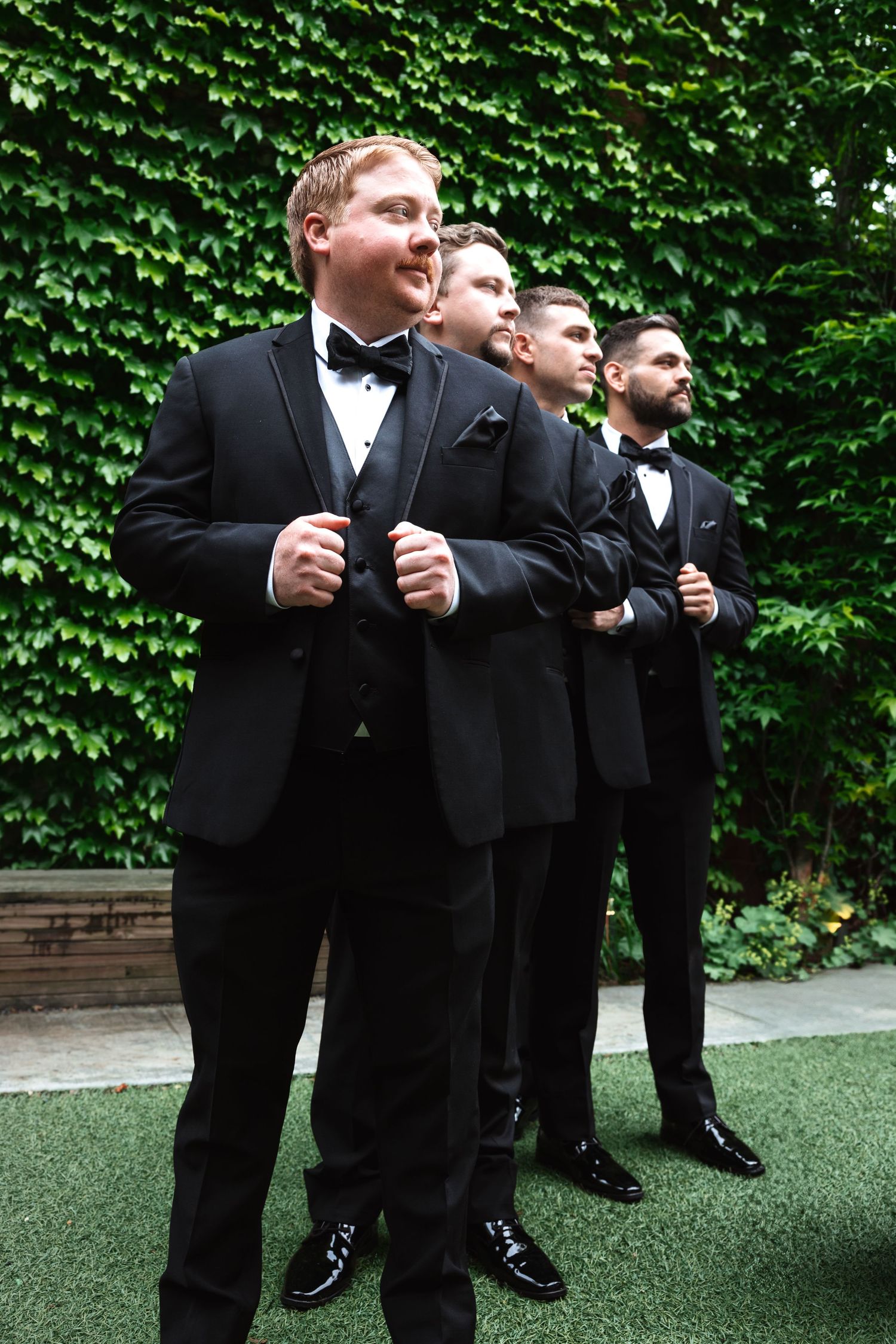 Four groomsmen in matching black tuxedos stand in a row outdoors against a lush green ivy backdrop.