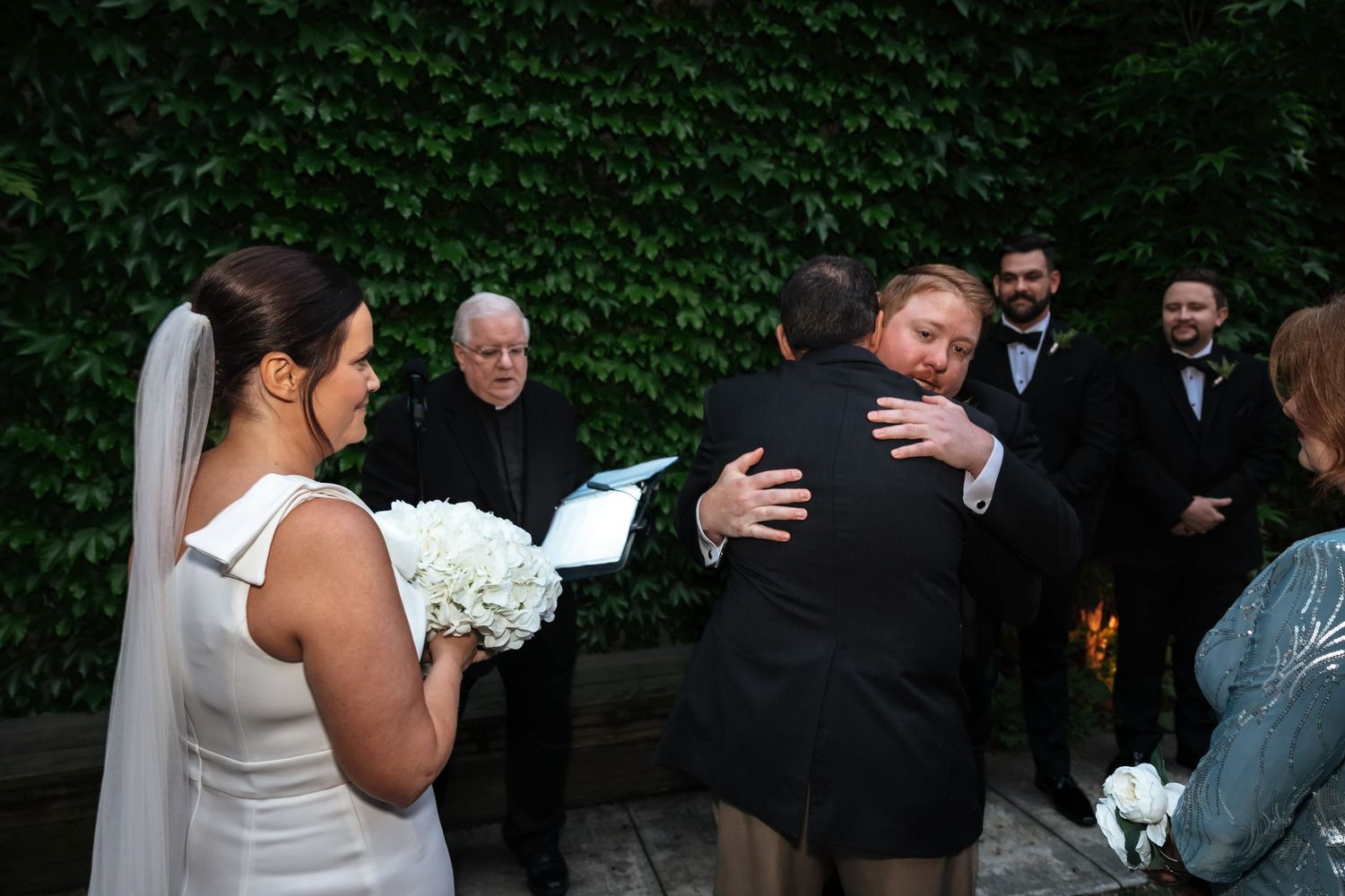 Bride watches as two guests embrace warmly during an outdoor wedding ceremony with ivy backdrop.