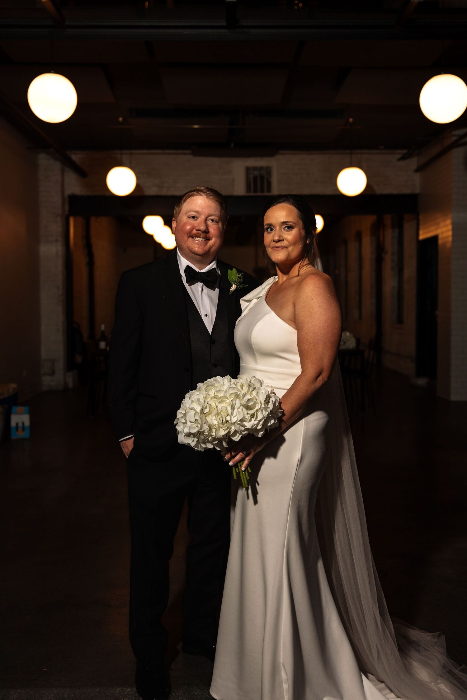 Happy wedding couple posing in industrial venue, bride holding white bouquet in sleek white gown.