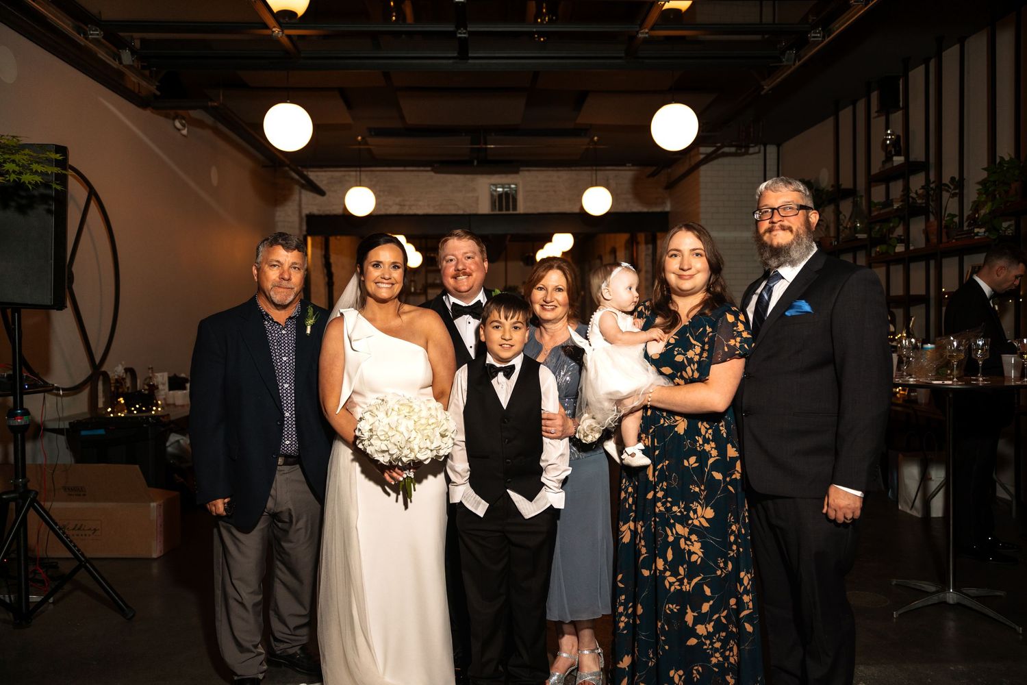 Wedding party group photo with bride in white gown, groom in tuxedo, and family members smiling in elegant venue.