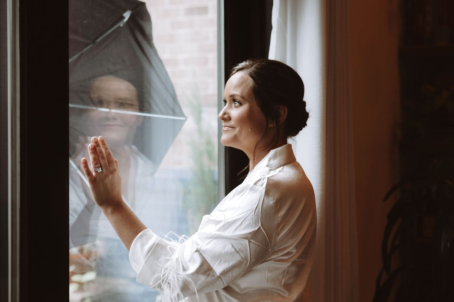 Smiling bride in white robe looking out window with her reflection in the glass.