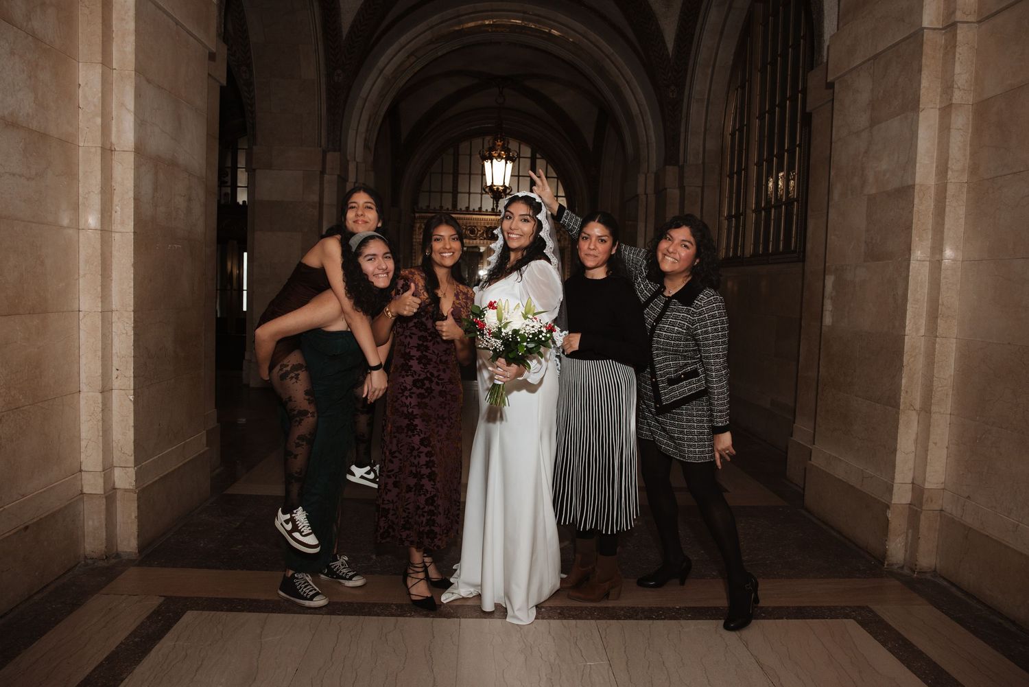 Bride surrounded by five smiling female guests in a grand stone corridor at her wedding.