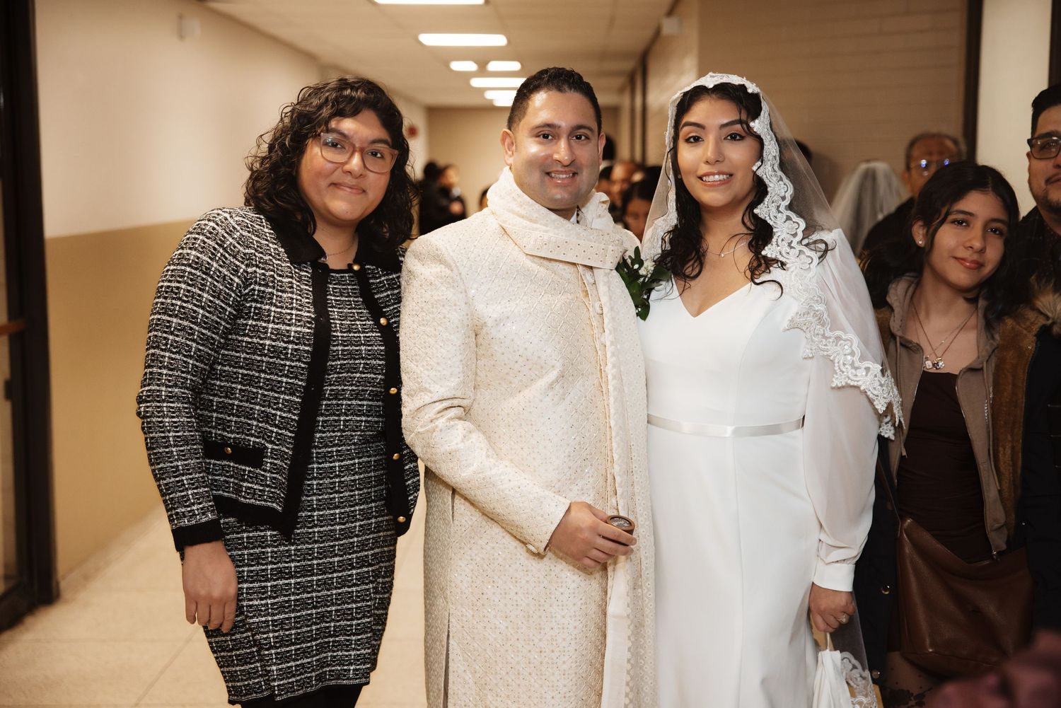 Bride in white veil and groom in ivory sherwani pose with two female guests in wedding venue hallway.
