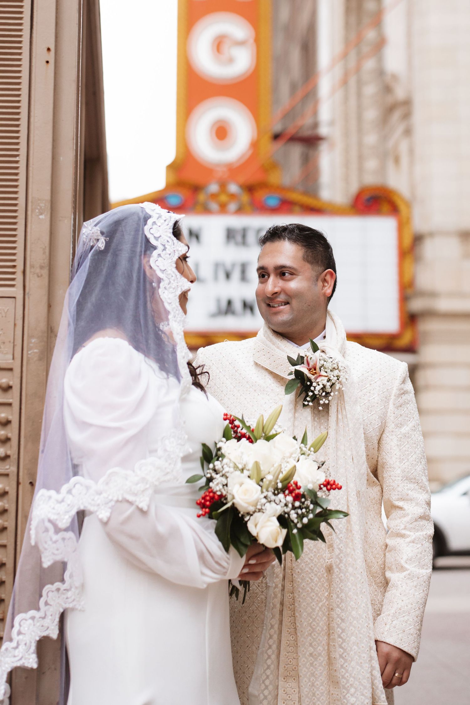 Groom in embroidered cream suit smiles at veiled bride holding white and red bouquet.