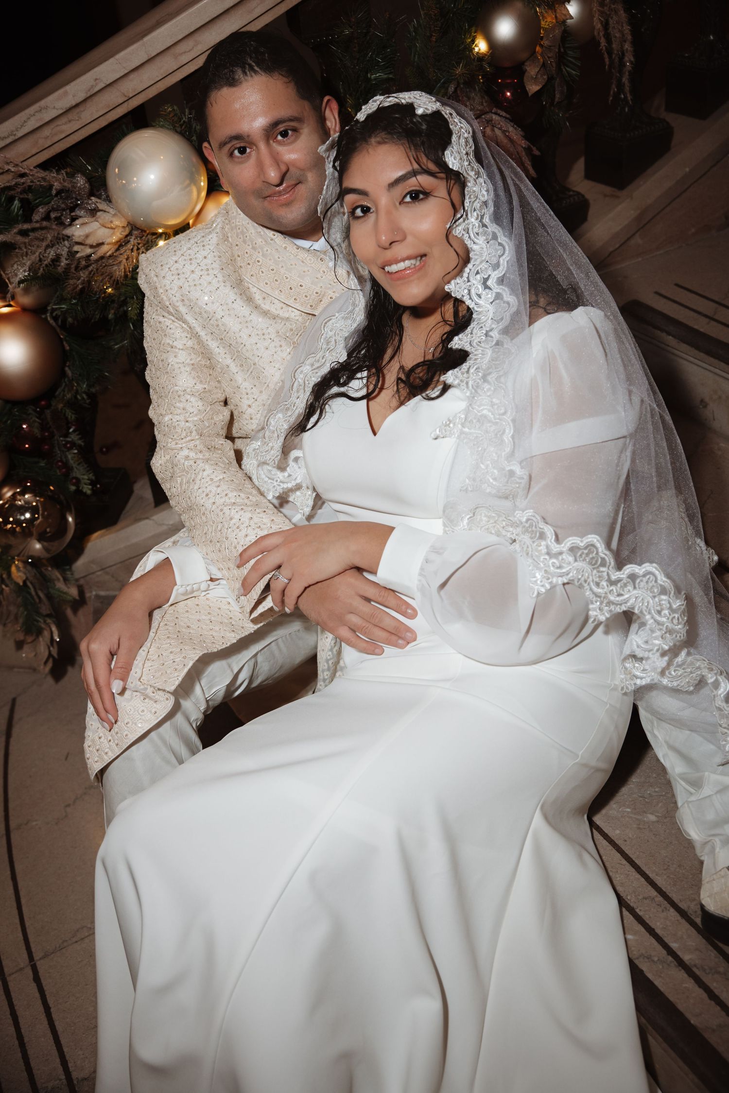 Close-up portrait of smiling bride in lace veil and groom in ivory suit on Christmas-decorated staircase.