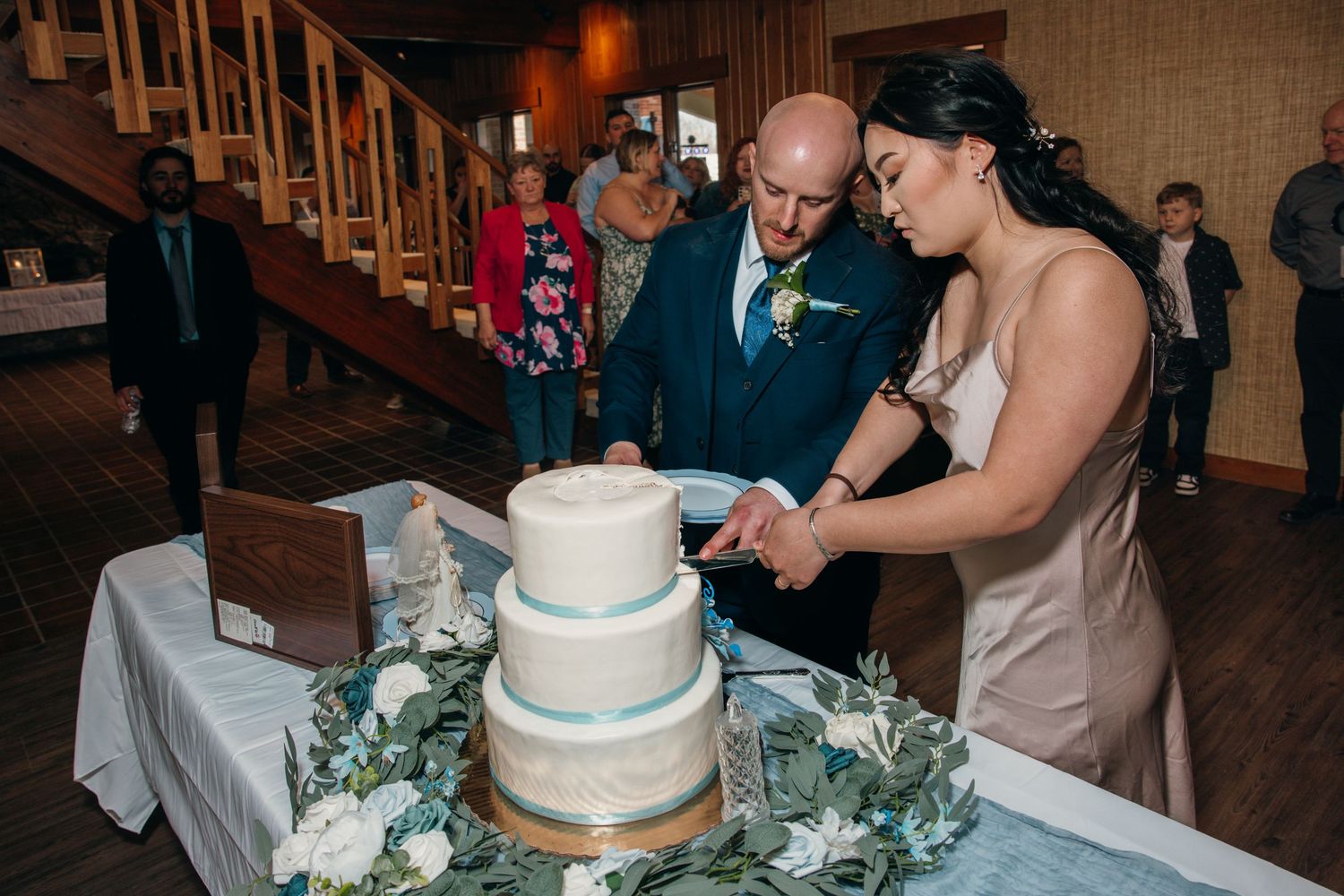Newlyweds cutting elegant three-tier white wedding cake decorated with blue accents and florals.