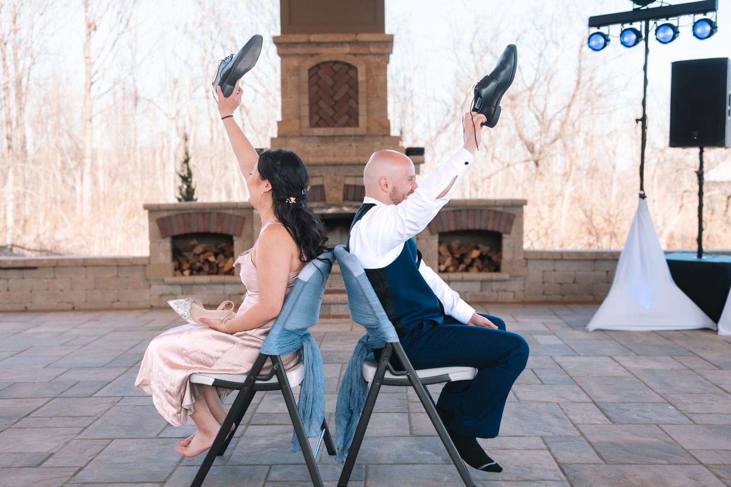 Bride and groom sit back-to-back on chairs raising shoes in a fun wedding game on an outdoor patio.