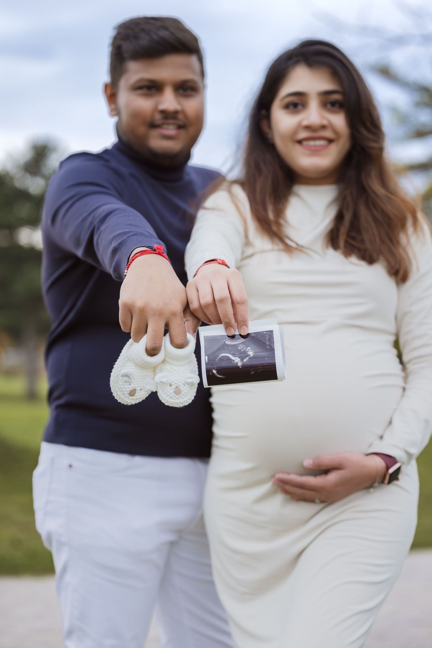Smiling couple showing baby shoes and ultrasound photo during outdoor maternity shoot.