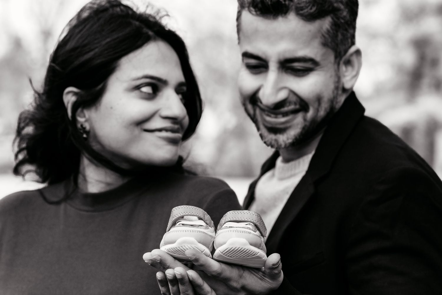 Black and white photo of happy expecting couple holding small baby sneakers outdoors.