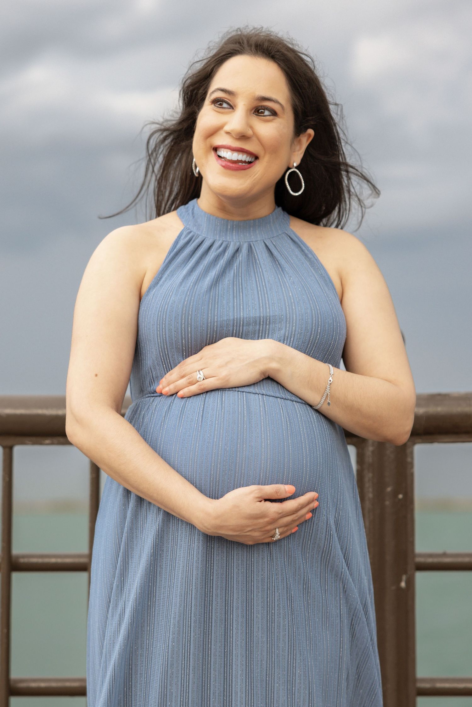 Smiling pregnant woman in blue halter maxi dress holding her baby bump on a waterfront pier with cloudy skies.