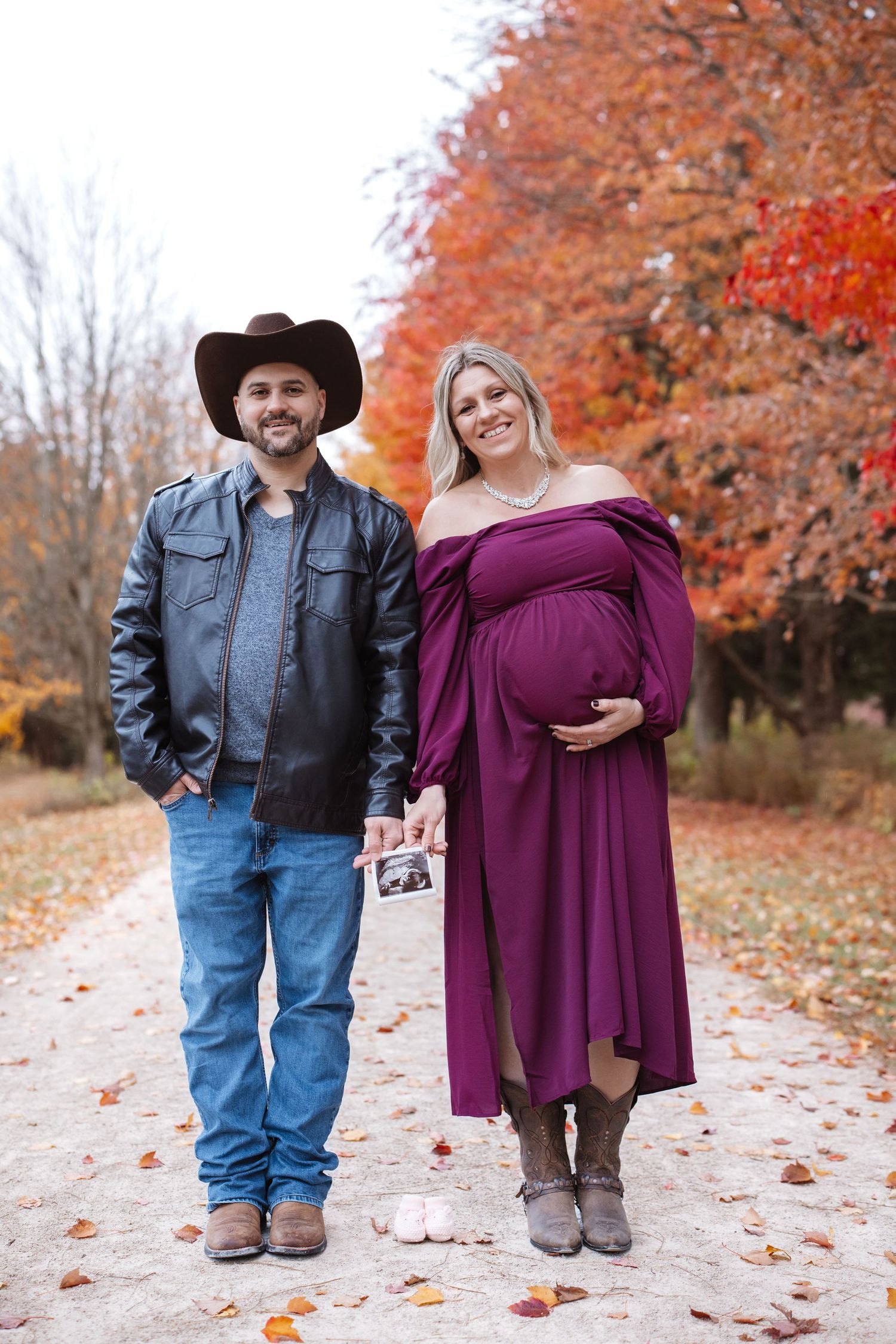 Smiling pregnant couple in autumn park holding ultrasound photo during fall maternity session.