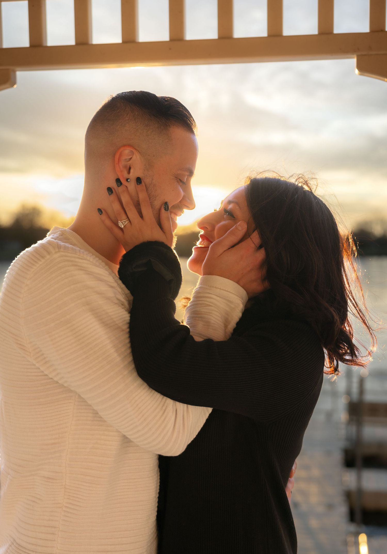 Couple sharing a romantic moment at sunset, woman smiling as man holds her face near a waterfront gazebo.