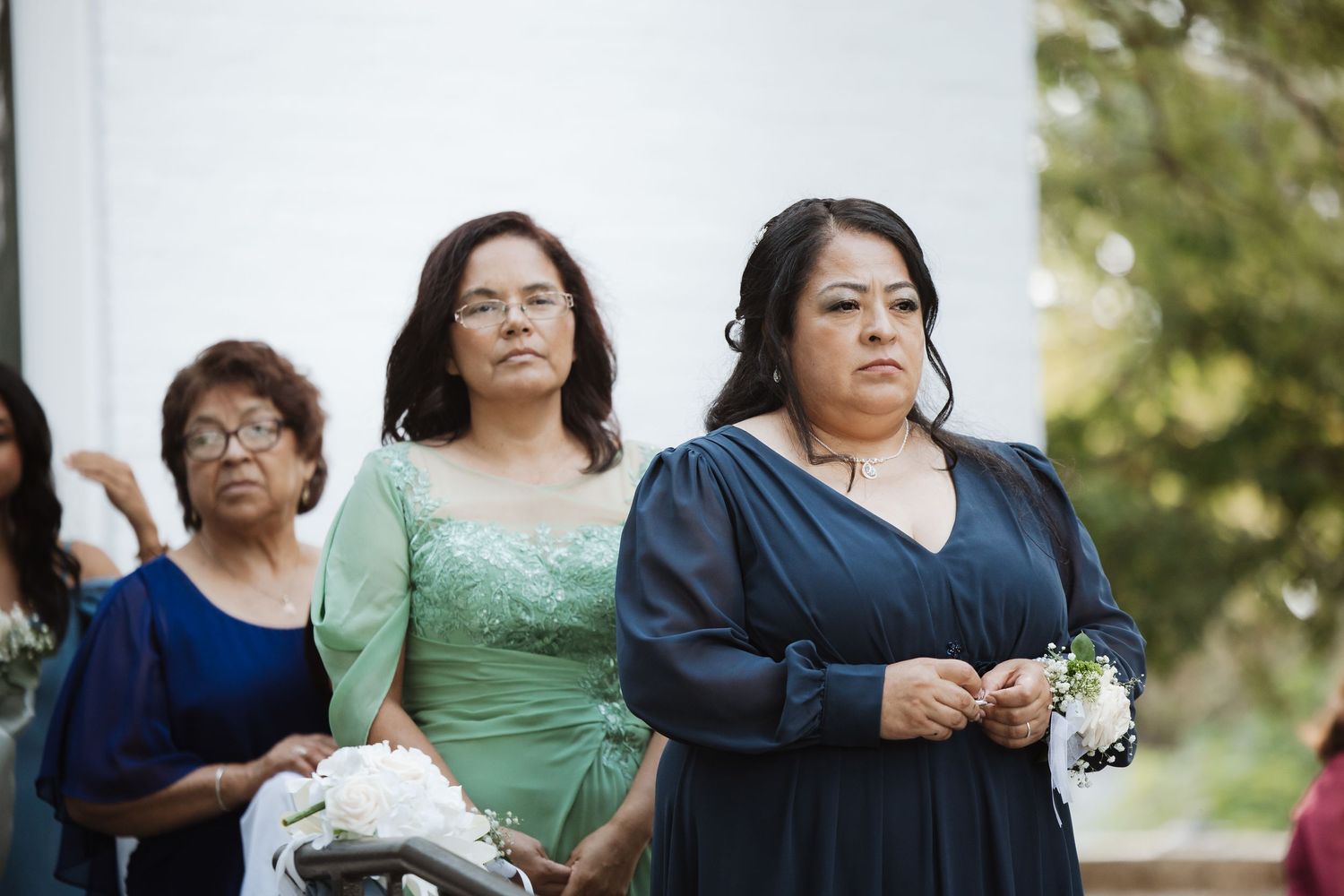 Women in green and navy formal dresses holding corsages at an outdoor wedding ceremony.